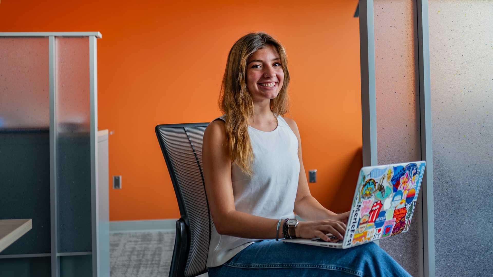 A blonde female student in a white top and denim skirt sits with a laptop on her knees in an office with gray cubicle walls in front of an orange wall.