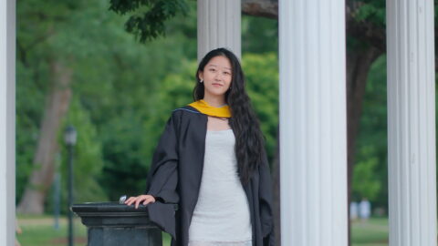 A student poses by the Old Well wearing master's degree graduation regalia.