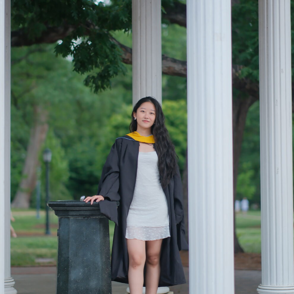A student poses by the Old Well wearing master's degree graduation regalia.